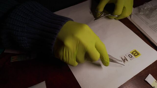 Sinister person assembles threatening message with tweezers and cut-out newspaper letters in dim setting.