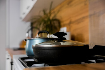 Close-up of a black frying pan with a transparent lid and a blue saucepan with a transparent lid and silicone pads on the handles against the background of a modern wooden-white kitchen out of focus