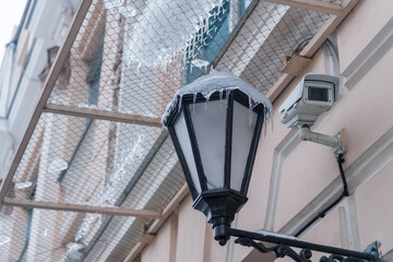 A decorative street lamp hangs from a building, draped in icicles, while a security camera observes the cold urban landscape. The scene captures the essence of winter.