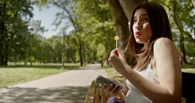 Young woman with lollipop and phone in park. Medium shot of woman with her phone while eating a lollipop.