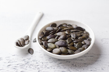 Raw pumpkin gymnosperm seeds in a white ceramic bowl with a spoon on a light blue background, healthy eating
