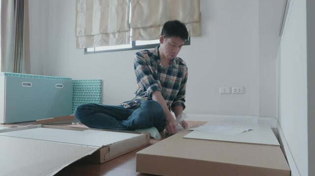 A young man sits on the floor, focused on assembling flat-pack furniture in a bright, minimalist room. The space features light colors and simple decor, emphasizing a tidy and modern aesthetic.