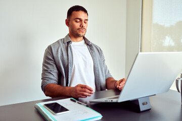 Young business man meditating at workplace with eyes closed, practicing mindfulness meditation during work on laptop