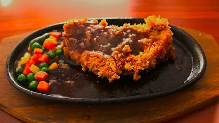 Crispy breaded chicken steak served on a hot hotplate, accompanied by fresh vegetables on the side. This dish offers a combination of savory flavors and interesting textures, perfect for food lovers. 