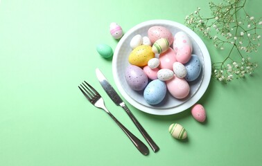 Easter decor concept. Top view photo of colorful eggs in a plate with knife and fork on pastel background
