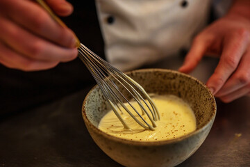 Chef expertly whisking creamy hollandaise sauce in a small bowl in a professional kitchen setting