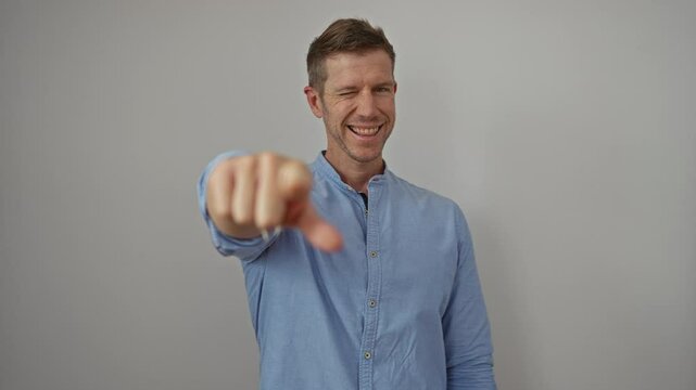 Young man pointing at camera confidently, smiling over white background, suggesting choice and involvement, wearing casual blue shirt, isolated and engaging