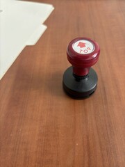 A close-up view of a red and black office stamp with a "Top" label placed on a wooden desk, accompanied by a file folder in the background. Suitable for office, work, and administrative themes