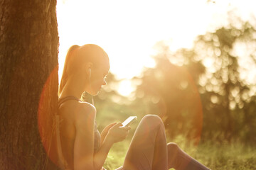 Young woman enjoying music on smartphone while sitting against a tree in golden sunlight, creating...