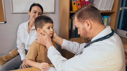 Male doctor inspecting young boy neck, lymph nodes with both hands during medical consultation. Attentive mother sitting in background. Concept of health care, medicine, treatment