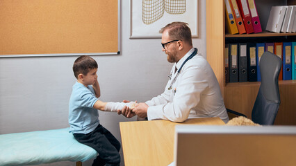 Young boy with bandaged arm visiting doctor for checkup and consultation., Bearded man carefully examining hand in his medical office. Concept of health care, medicine, treatment