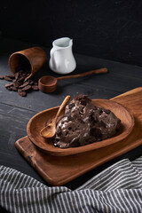 Indulge in creamy chocolate ice cream presented on a wooden platter, accompanied by a wooden spoon and cacao nibs on a rustic table