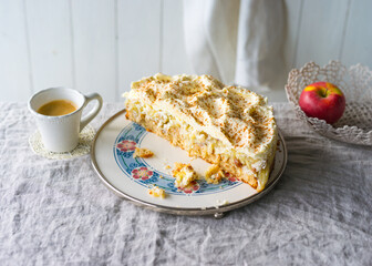 A slice of creamy apple cake is presented on a vintage plate next to a cup of coffee and a bright red apple. The soft, rustic tablecloth adds a warm touch to the arrangement