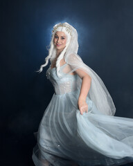 Close up portrait beautiful blonde fantasy maiden faery wearing medieval fairytale ballgown costume, veiled white cape. standing pose isolated dark studio background with mystical smoke fog effect