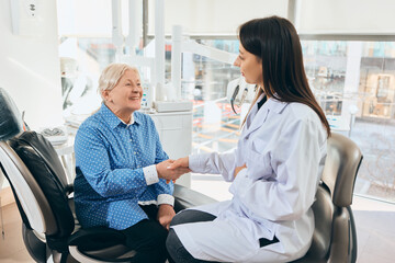 Fototapeta premium Elderly woman seated in dental chair shaking hands with female doctor in lab coat, smiling, medical tools and equipment visible in bright room. Concept of healthcare and pensioners, medicine.