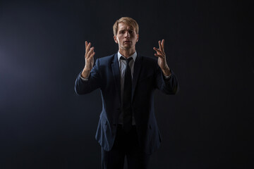 In a dimly lit studio, a man in a dark suit and tie gestures expressively. The sleek attire and...