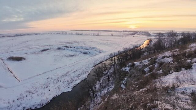 As winter progresses, a sunset casts warm hues over a frozen river valley, surrounded by serene, snowcovered fields and tall trees, creating a tranquil, picturesque scene