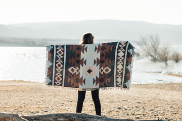 Woman holding a colorful blanket beside the water at a serene lakeside during sunset