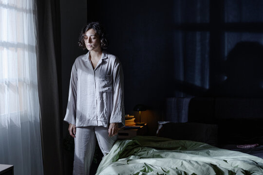 Wide shot of woman with closed eyes dressed in pajamas standing near window silently in bedroom