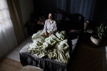 High angle shot of adult woman suffering from sleepwalking sitting in middle of bed while moonlight...
