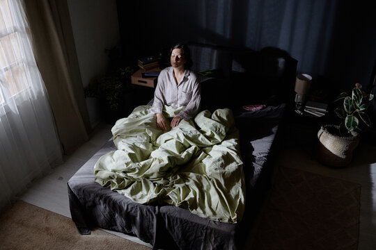 High angle shot of adult woman suffering from sleepwalking sitting in middle of bed while moonlight illuminating her