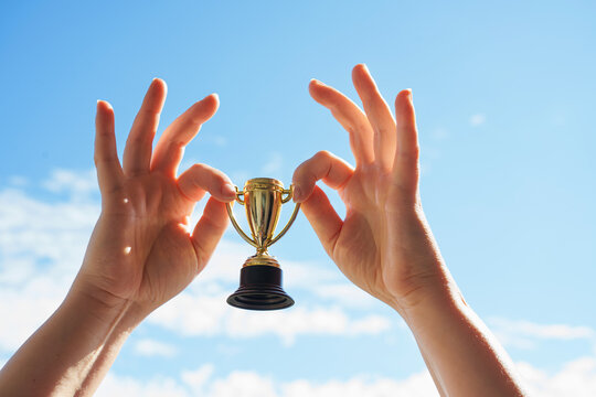 Hand holds and raised small cup trophy, holding cup against clean sky backgrund. Consolation prize award and victory concept.