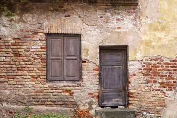 
A fragment of an old building with decayed plaster, exposing a brick wall with a wooden door and a window with closed sashes.