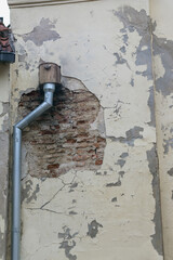 
A small hole in the wall of an old building with peeling plaster is visible, in which a birdcage is installed and a rainwater pipe is attached.