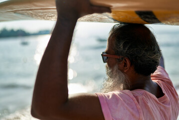 Senior man with surfboard walk along ocean shore. Elderly surfer enjoys active lifestyle by sea....