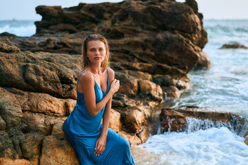 Elegant woman in wet dress sits by ocean rocks on tropical island. Waves crash. Model poses by sea...