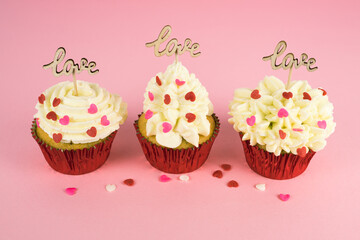 Valentine's Day. Three cupcakes with cream and red topping decoration with a wooden sign with the word LOVE on a pink background. Copy space.