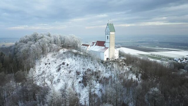 Drone footage of a snowy winter landscape as it moves away from a hilltop church, revealing a serene snow-covered forest in the foreground. A peaceful and cinematic winter scene.