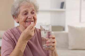 Smiling senior woman with glass of water taking pill at home. Space for text