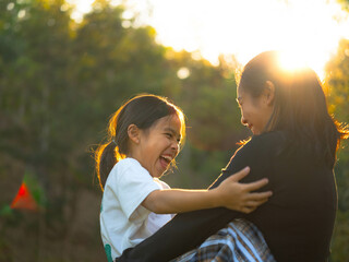 Fototapeta premium Mother and daughter are playing together in the park. Happy mother hugging her daughter sitting in the park with the sunset light in nature.