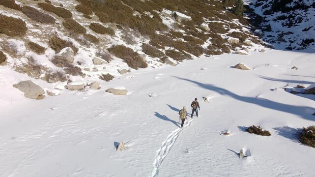 Two tourists with backpacks walk along a snowy valley in the mountains, trampling a path. Aerial view of two tourists with backpacks in the mountains in winter.