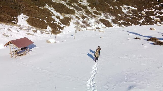 Two tourists with backpacks walk along a snowy valley in the mountains, trampling a path. Aerial view of two tourists with backpacks in the mountains in winter.