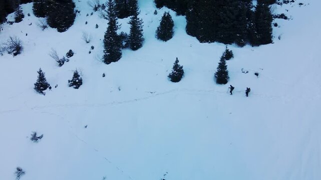 Two tourists with backpacks walk along a snowy valley in the mountains, trampling a path. Aerial view of two tourists with backpacks in the mountains in winter.