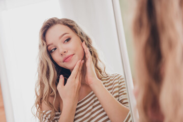 Charming young woman with blond hair gazing at herself in mirror, reflecting self-care and happiness, dressed in a striped pullover, enjoying leisure time indoors in a cozy living room setting.