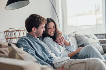 Young happy couple having fun talking laughing relaxing at home on couch, boyfriend embracing girlfriend telling funny joke sitting on sofa, humor in relationships, enjoying weekend together.