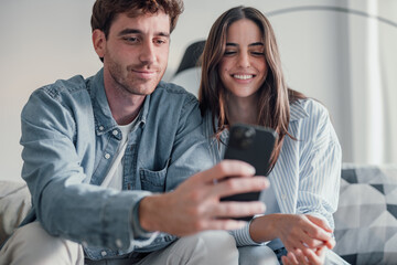 Happy millennial couple sit relax on couch in living room watching video on cellphone together, smiling young husband and wife rest on sofa at home browsing Internet using modern phone device.