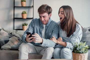 Excited millennial husband and wife relax on couch in kitchen feel euphoric win lottery online on...