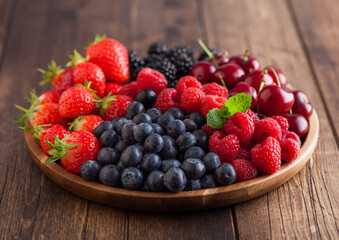 Fresh organic summer berries mix in round wooden tray on light wooden table background. Raspberries, strawberries, blueberries, blackberries and cherries. Macro