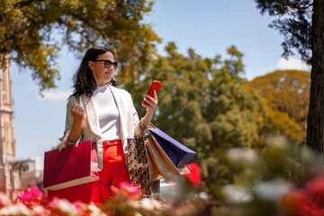 mulher de 50 anos passeando no parque com sacolas de compras e celular na mão