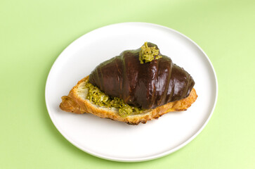 Croissant stuffed with pistachio paste and kadaif dough covered with dark chocolate on a gray plate on a green background. Unusual pastry with a delicate filling. Trend food concept. Selective focus.