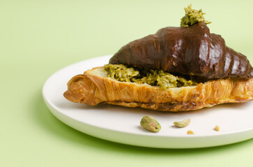 Croissant stuffed with pistachio paste and kadaif dough covered with dark chocolate on a gray plate on a green background. Unusual pastry with a delicate filling. Selective focus. Copy space