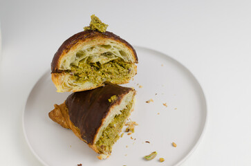 Croissant stuffed with pistachio paste and kadaif dough covered with dark chocolate on a gray plate in the cut. Unusual pastry with delicate filling. Trend food concept. Selective focus. Copy space.