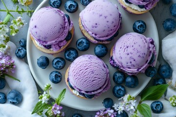 Blueberry ice cream scoops on small cookies forming mini sandwiches garnished with fresh blueberries and flowers