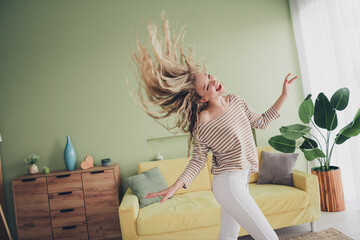 Young woman joyfully dancing in bright living room with stylish decor, expressing happiness and freedom on a relaxing weekend at home, wearing a striped pullover and enjoying daylight indoors.