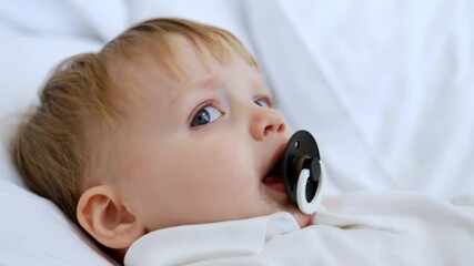 Cute Caucasian baby boy with fair hair lies on the pillow. Kid with pacifier is falling asleep. Close up.