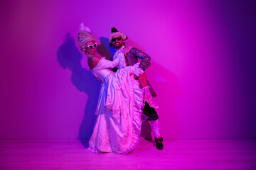 Elegant couple in royal-themed costumes and sunglasses dancing under pink neon lights